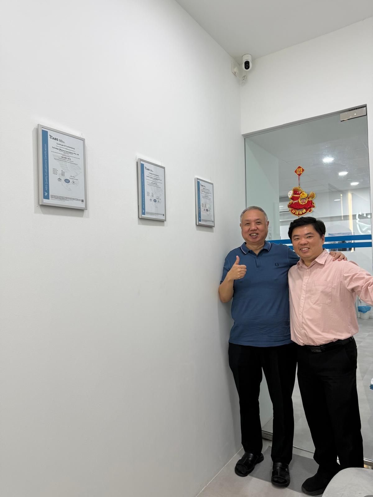 Two Techwise team members smiling beside the framed ABS ISO certificates in the office