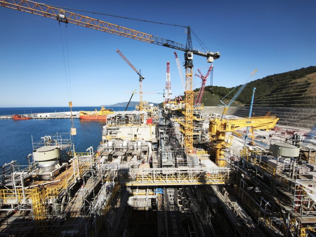 Floating production unit topsides under conversion at a shipyard quayside, with tower cranes overhead